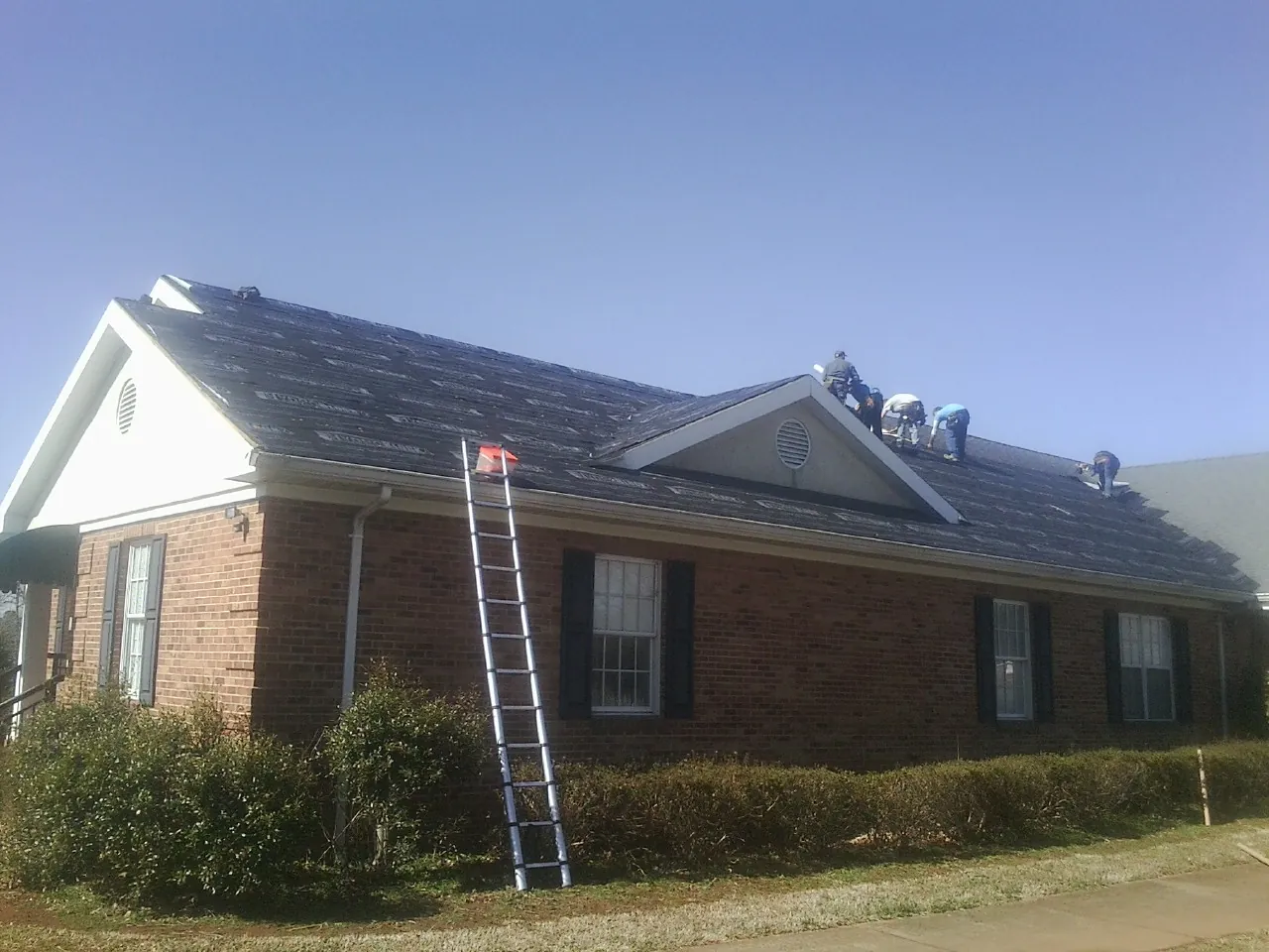 Crew tearing shingles off a brick ranch roof with a ladder set — Emergency Roof Repair work by our crew in Rexford