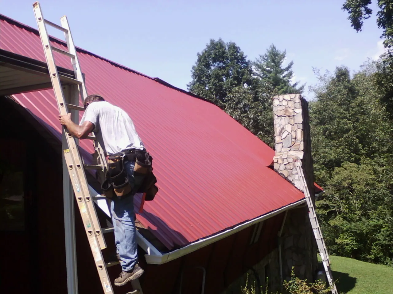 Roofer on a ladder beside a red metal roof and stone chimney — Soffit & Fascia Repair by Roofing Brothers in Rexford