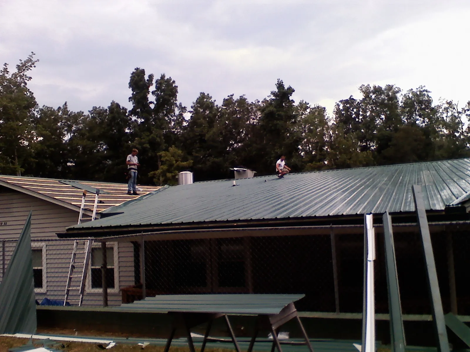 Two roofers installing green metal panels above a covered porch in Rexford